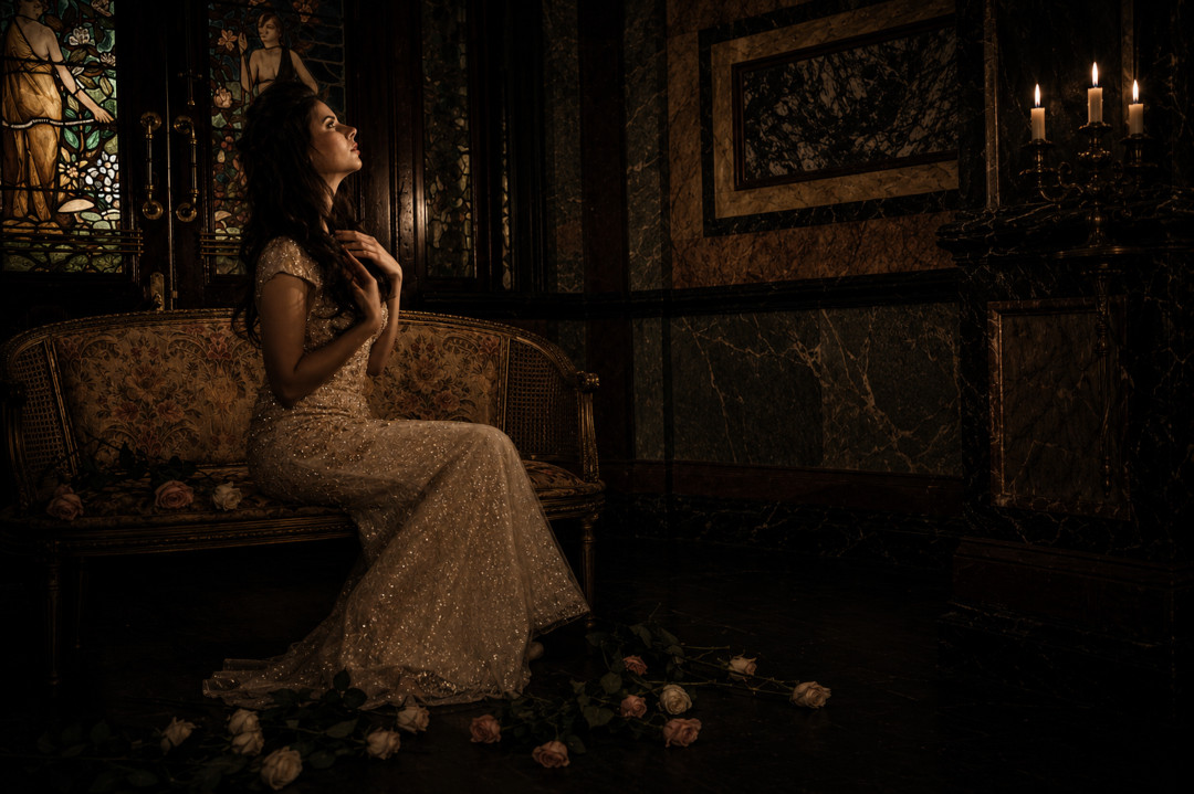 A bride seated in candlelight within a marble and stained-glass interior, roses resting on the floor.