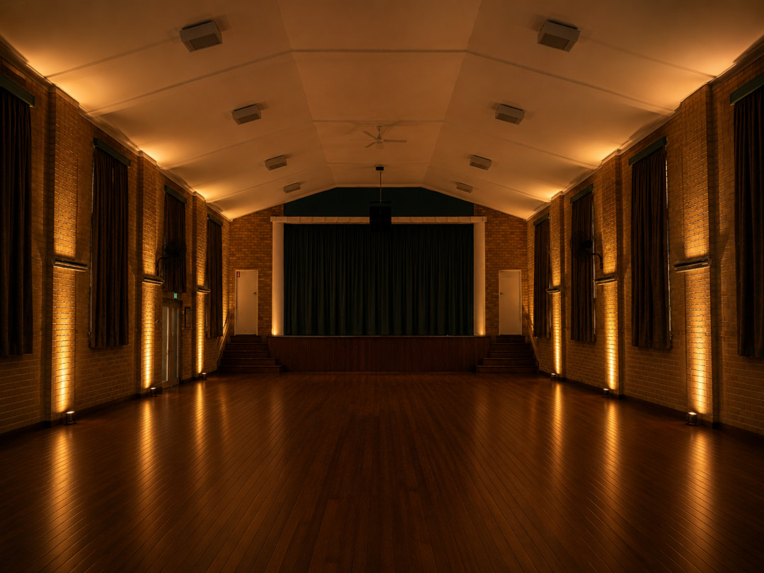The Parish Hall interior, empty and warmly lit by uplights along the brick walls, with polished wooden floors and a stage at the far end